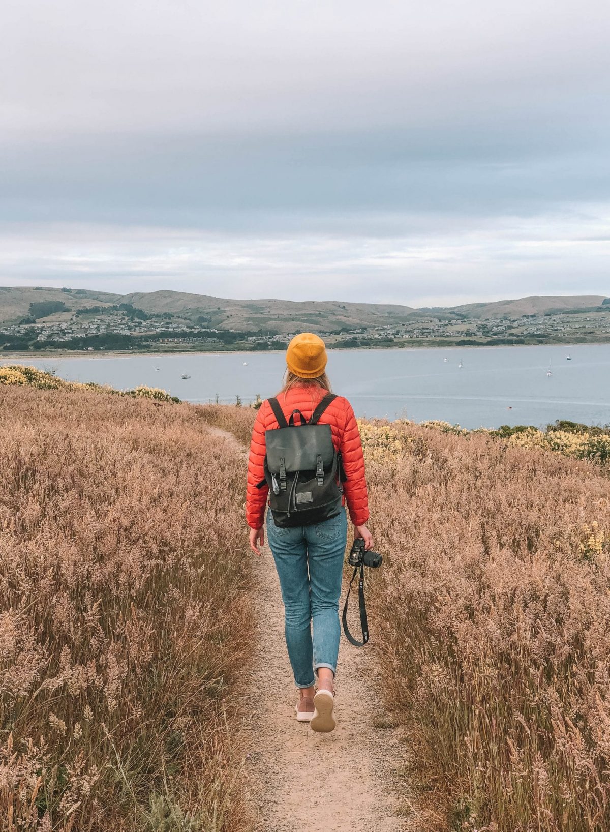 An outdoorsy woman walking on a hiking path with a backpack and camera in hand, looking out towards the water and hills in the background--outdoor gifts for women.