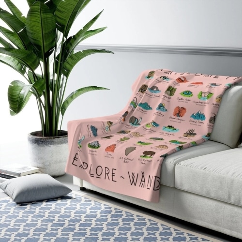 The pink National Parks Sherpa Blanket laying across a grey couch with a plant in the background and a pillow and carpet on the ground.