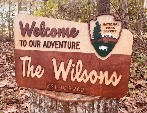 A wooden National Park Service Custom Welcome Sign that says Welcome to Our Adventure and The Wilsons, sitting on a tree stump outdoors.