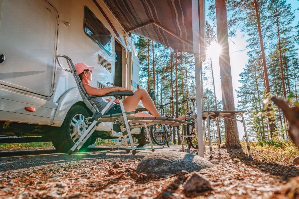 Woman lounging on a camp chair at her campsite next to her RV, looking out toward the trees. Best Gifts for RV Owners featured image.