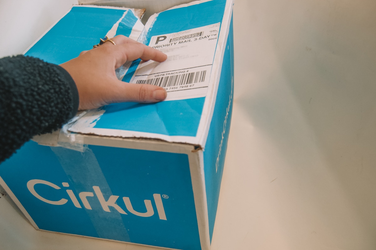 A hand sitting on top of a blue Cirkul box with postage visible.
