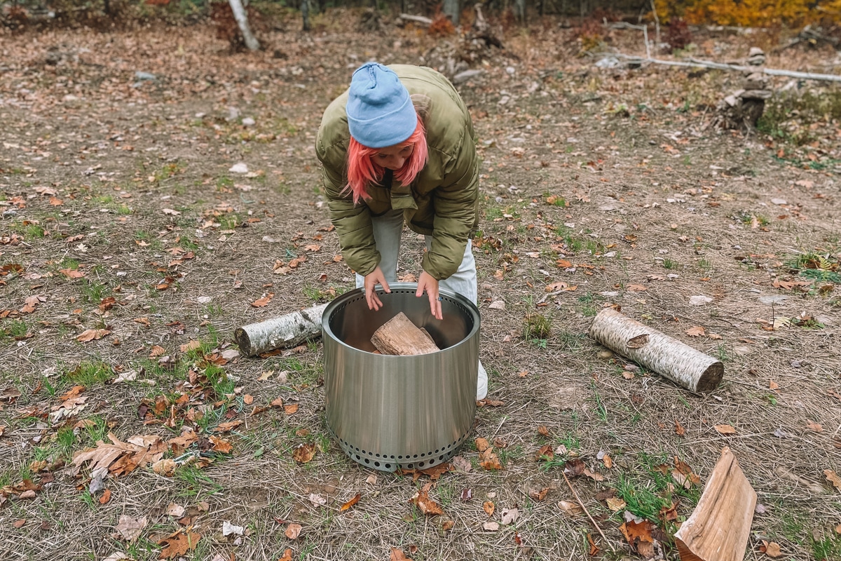 Woman with pink hair bent over a Solo Stove, placing a piece of firewood inside.