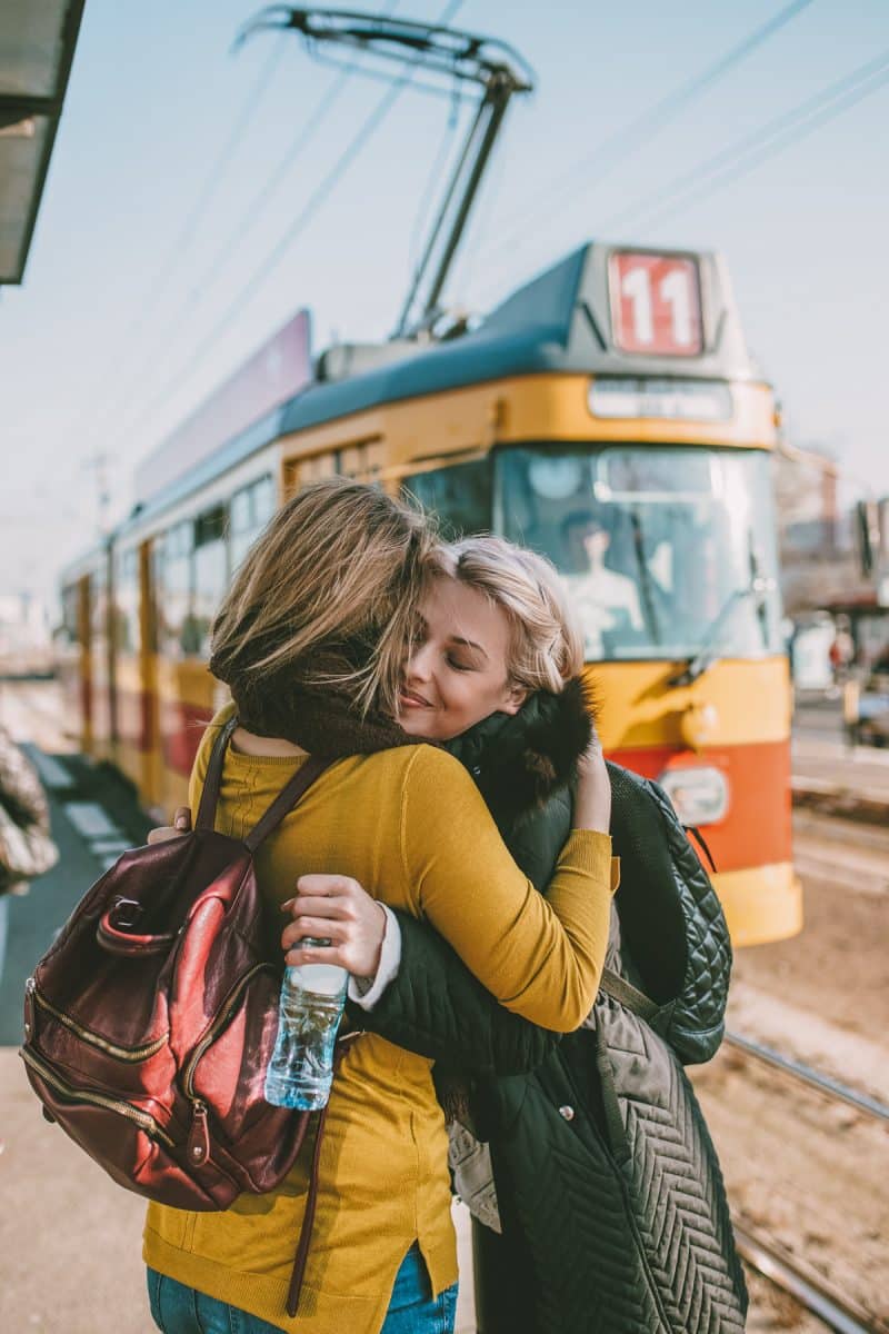 Two female friends smiling and hugging at a tram station with a yellow tram in the background.