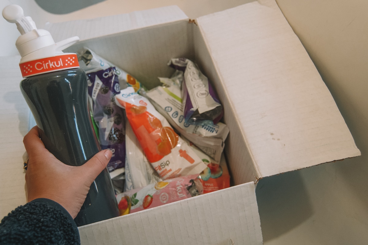 A hand pulling a Cirkul water bottle out of a box full of flavor cartridges in their packages.