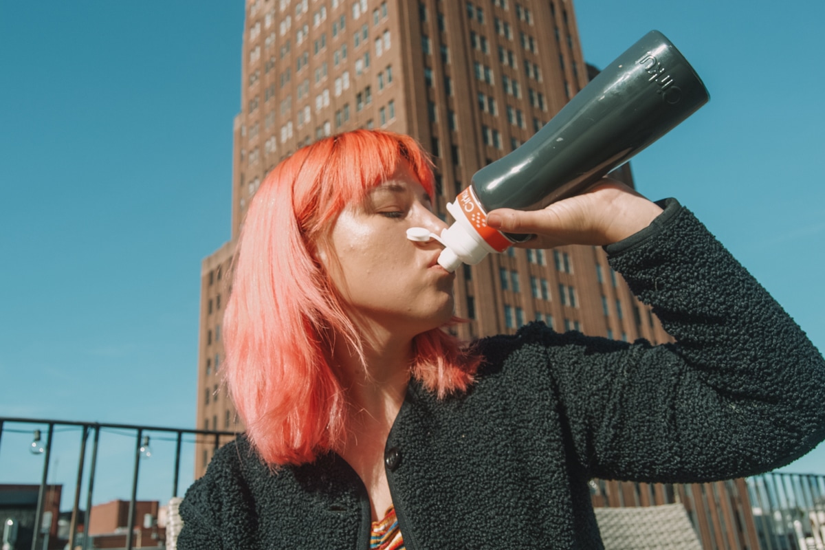 A woman with pink hair in profile taking a sip from a  Cirkul water bottle with a blue sky and tall building in the background.