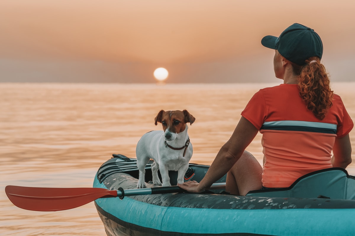 A woman in an orange shirt with a curly ponytail faces away while sitting in a blue kayak at sunset, with a small terrier in the boat.