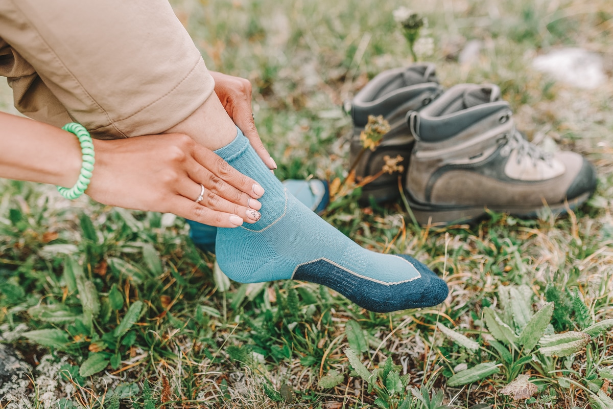A woman's hands pulling up her blue hiking socks; she's wearing khaki pants and a diamond wedding ring, and a pair of hiking boots sit on the grass beside her.