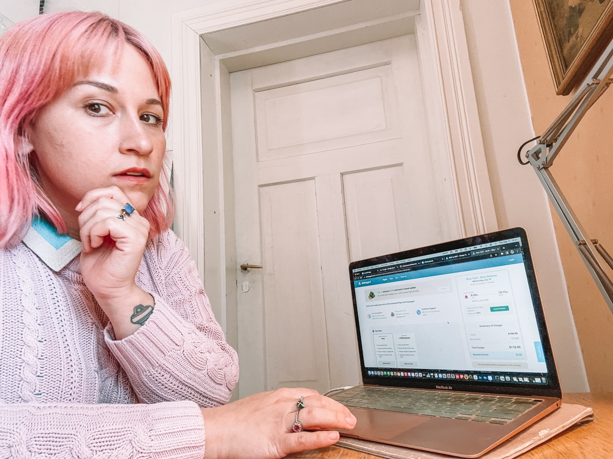 A woman with pink hair and a pink sweater sits at a desk and looks at the camera thoughtfully, with the  Skiplagged website open on her laptop and a closed white door in the background.