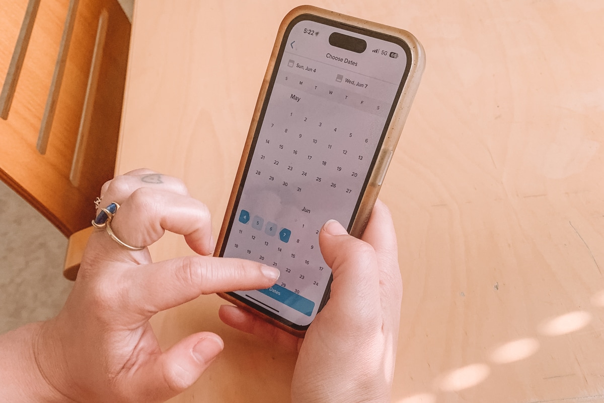 A pair of hands holding an iPhone, choosing dates on the Hopper app while resting on a light wood kitchen table.