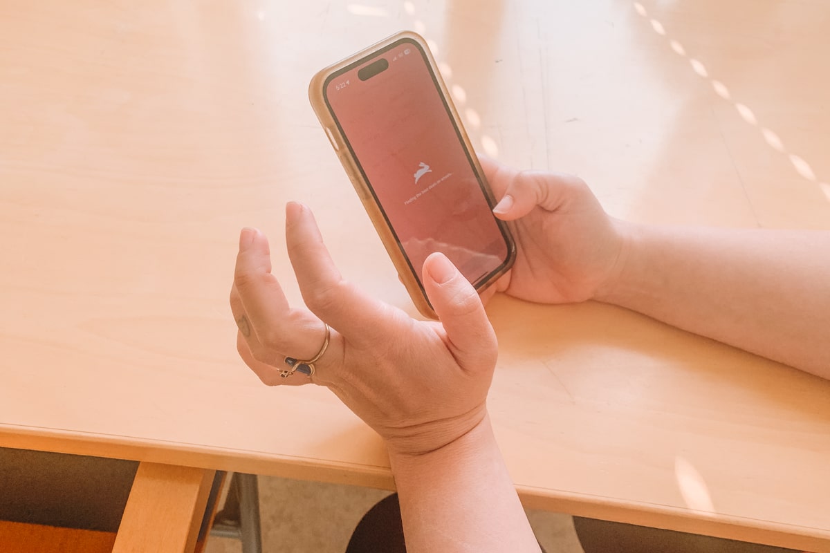 A pair of hands holding an iPhone, browsing on the Hopper app while resting on a light wood kitchen table.