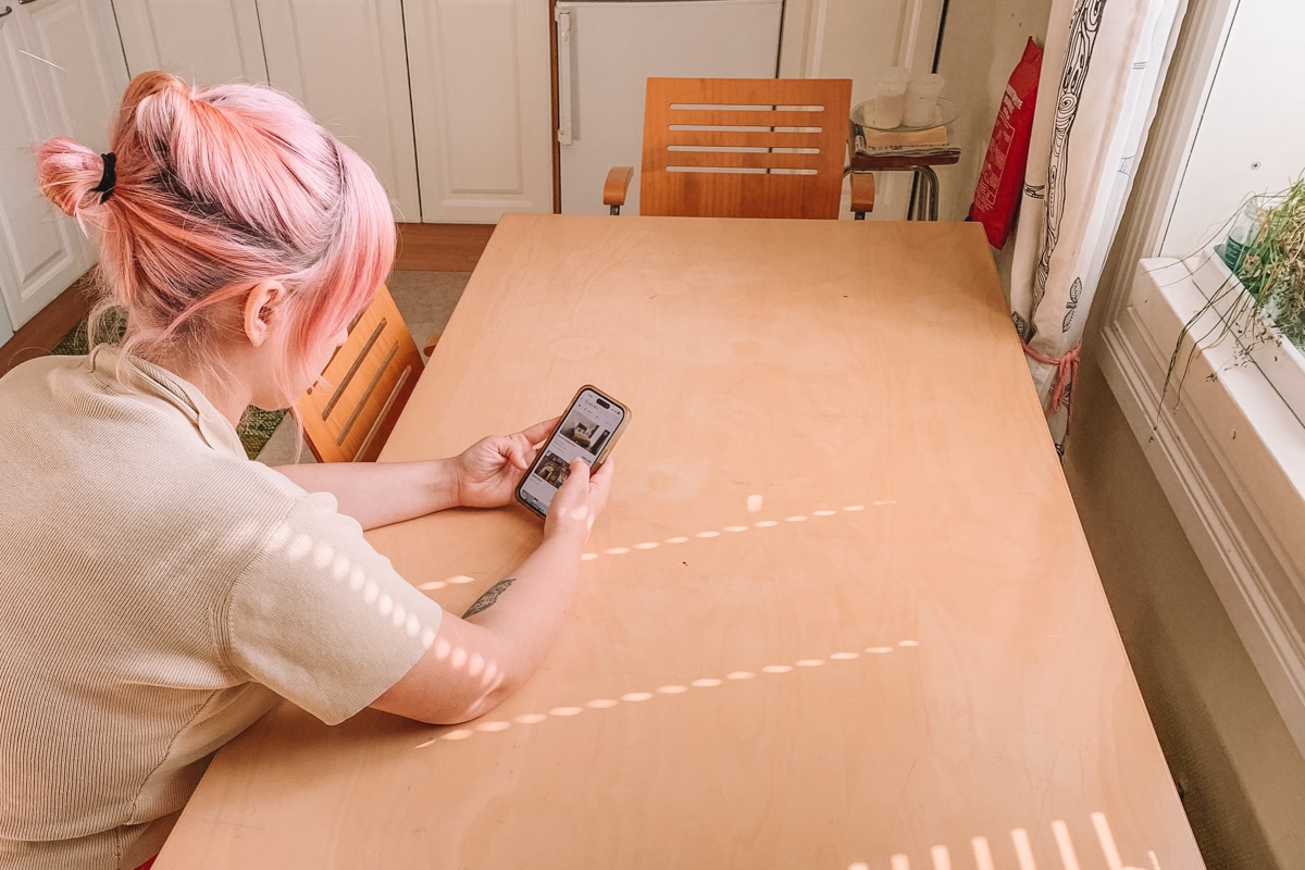 A young woman with light pink hair holding an iPhone, browsing hotels on the Hopper app while resting on a light wood kitchen table.