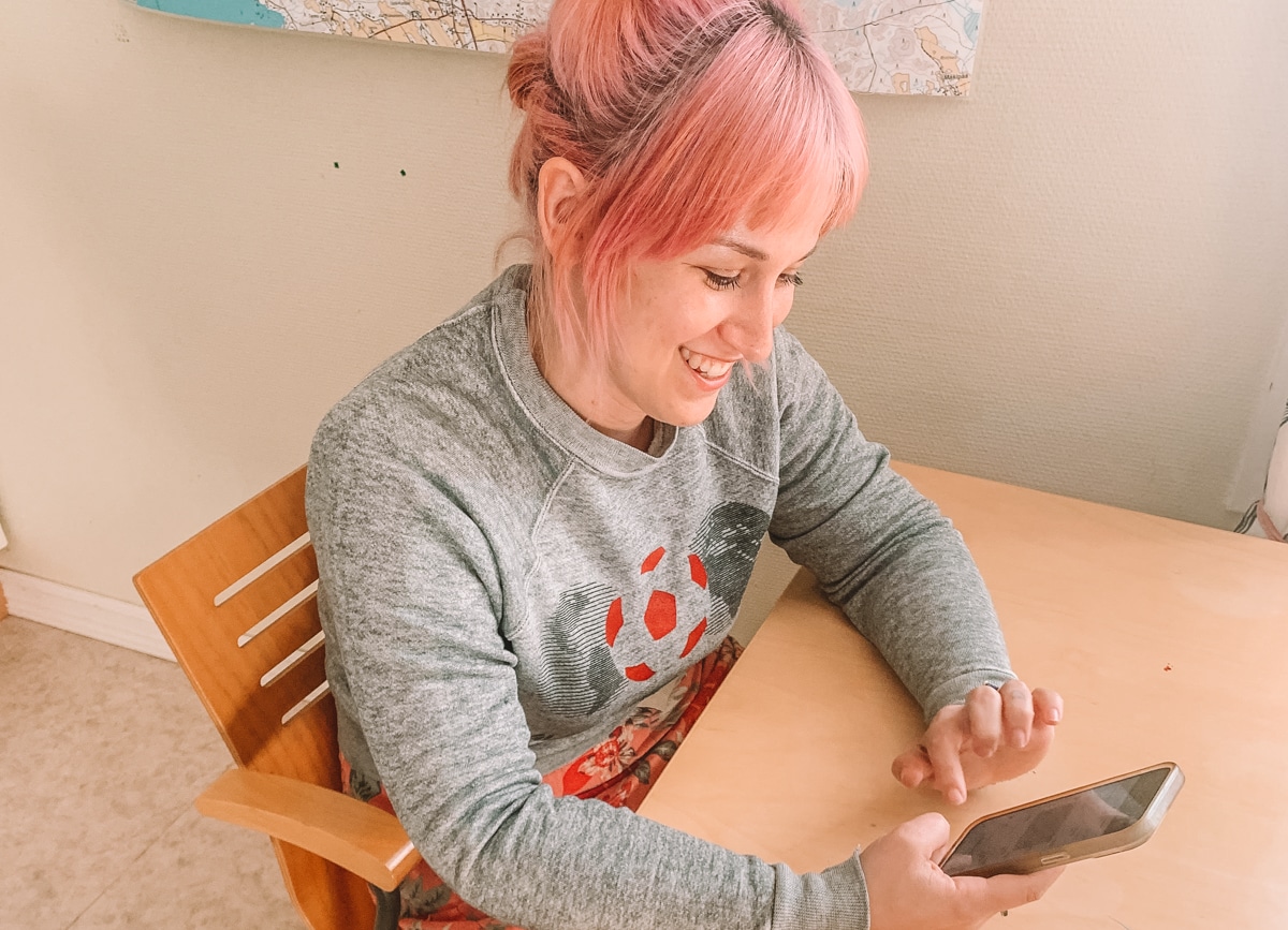 A woman with pink hair and a grey sweatshirt grins as she sits at a light wood kitchen table and looks at the Skiplagged app on her iPhone.