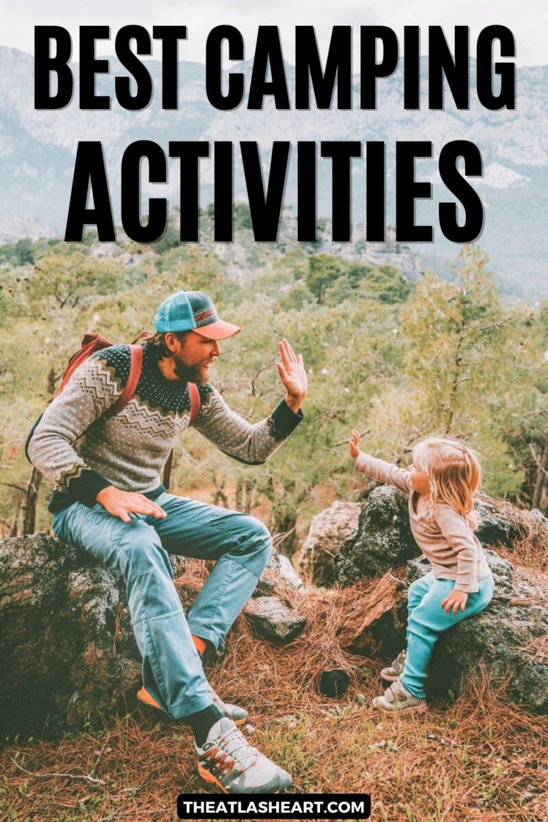 A man in a patterned sweater and a blue and orange baseball cap high-fives a blond toddler as they sit on rocks covered in pine-needles overlooking a forested mountain vista, "Best Camping Activities."