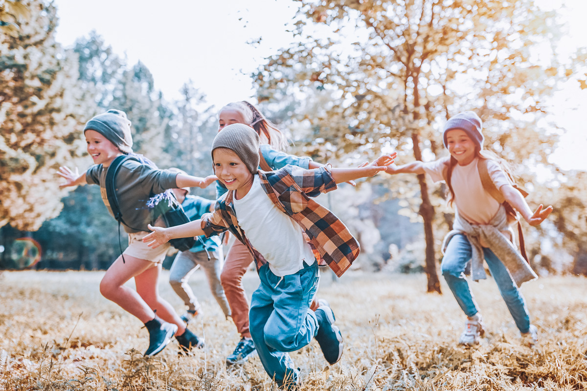 A group of four children wearing knitted beanies running across a grassy field with trees in a soft focus in the background.