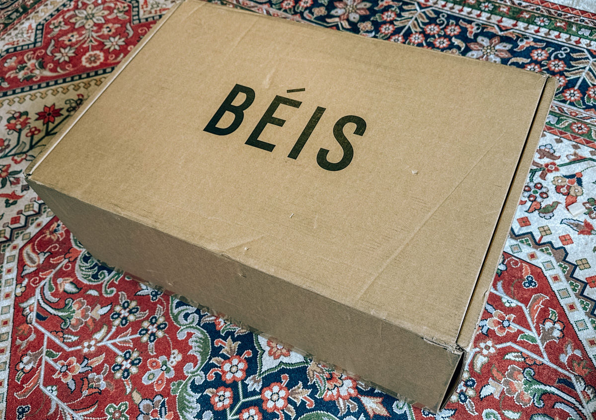 A cardboard box with "Beis" printed on it, sitting on a red, blue, and white oriental rug. 