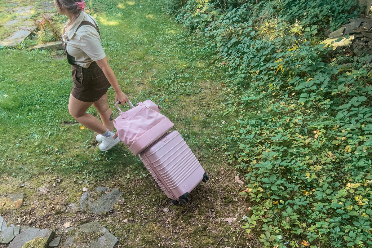 A woman in brown overall shorts wheeling a pink Beis suitcase, with a Beis Mini Weekender bag on top of it walking across a green lawn.