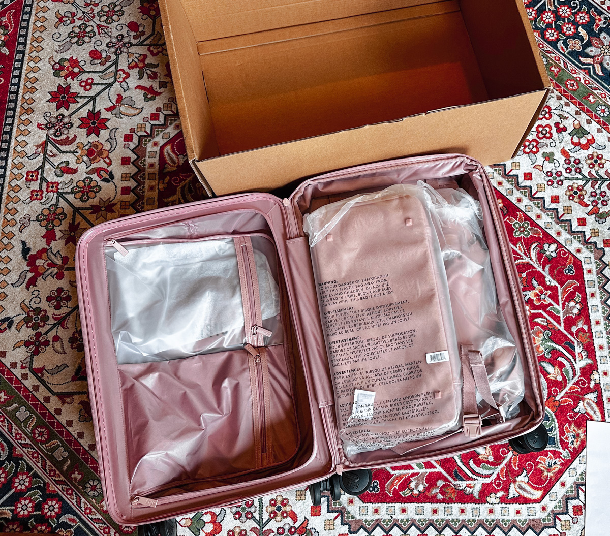 A new pink Beis suitcase laying open  on an oriental rug, with the carboard box it came in visible in the background. 